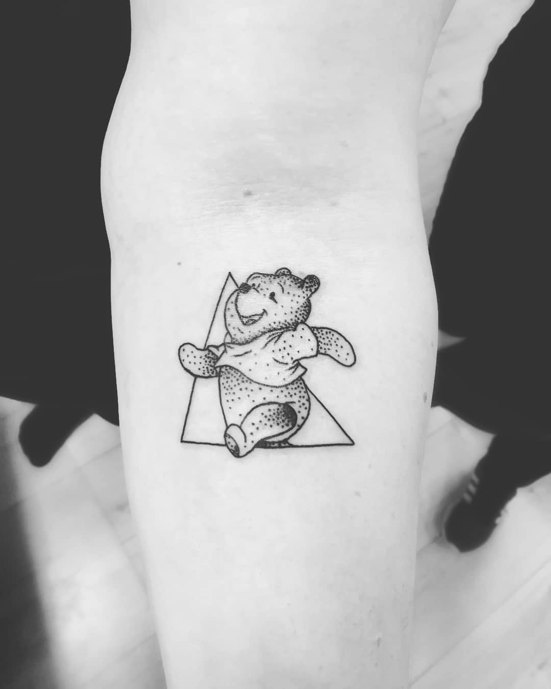 Black and white dotwork tattoo of Winnie the Pooh happily walking in front of a triangle outline on a person's arm
